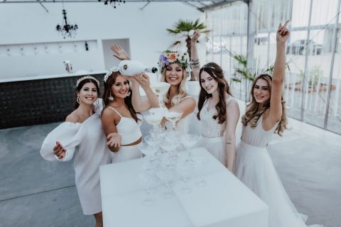 Hochzeitswahn - Fünf Frauen in weißen Kleidern feiern auf einer Party. Eine gießt Champagner in einen Turm aus Gläsern, eine andere trägt einen Blumenkranz. Sie alle lächeln fröhlich. Die Party findet in einem modernen, luftigen Veranstaltungsort mit einer Bar im Hintergrund statt.