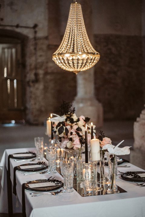Hochzeitswahn - Elegantes Speisezimmer in einem schwach beleuchteten Saal mit einem langen, schwarz-weiß gedeckten Tisch, geschmückt mit Kerzen, klarem Glas und einem Blumengesteck unter einem großen Kronleuchter. Innviertler-Versailles