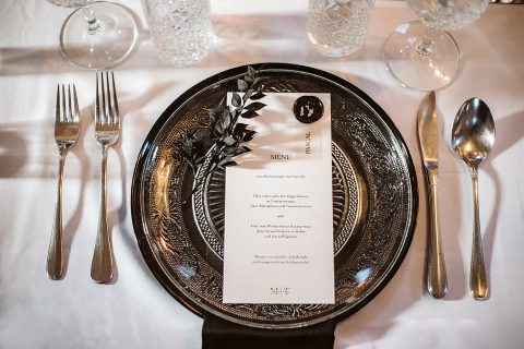 Hochzeitswahn - Ein elegantes Abendessen mit einem silbernen Platzteller, einer detaillierten Menükarte, Besteck und Gläsern auf einer weißen Tischdecke, dekoriert mit einem kleinen Olivenzweig. Innviertler-Versailles