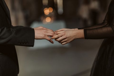 Hochzeitswahn - Eine Nahaufnahme der Hände eines Mannes und einer Frau, während der Mann der Frau einen Ehering an den Finger steckt, vor einem sanft verschwommenen Hintergrund mit warmer Beleuchtung. Innviertler-Versailles