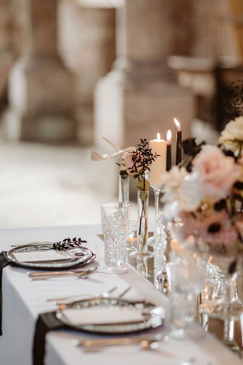 Hochzeitswahn - Elegante Tischdekoration für eine formelle Veranstaltung mit brennenden Kerzen, Blumenarrangements, feinem Porzellan, Gläsern und Silberbesteck, alles arrangiert auf einer weißen Tischdecke in einem sanft beleuchteten Veranstaltungsort. Innviertler-Versailles