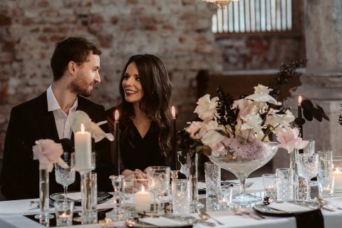 Hochzeitswahn - Ein Paar lächelt und unterhält sich bei einem romantischen Abendessen mit Kerzen und einem großen Blumengesteck in einem schwach beleuchteten rustikalen Raum. Innviertler-Versailles
