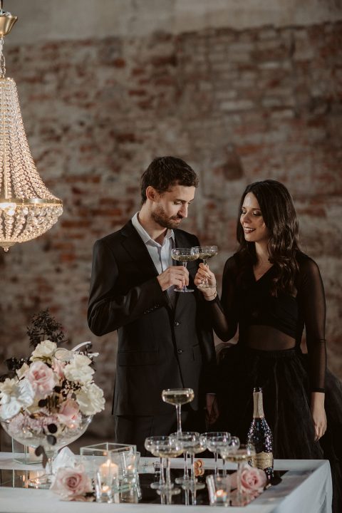 Hochzeitswahn - Ein Mann und eine Frau in Abendgarderobe stoßen mit Weingläsern an einem Tisch bei Kerzenlicht an, umgeben von eleganten Blumenarrangements und einem Kronleuchter in einer rustikalen Umgebung mit Backsteinmauern. Innviertler-Versailles