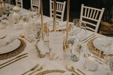 Hochzeitswahn - Elegante Tischdekoration für einen Hochzeitsempfang in den Farben Weiß und Gold, mit Kerzen, Blumenschmuck und einem Menü mit dem Titel „Lasst uns essen!“ auf jedem Teller. Der Tisch ist von weißen Stühlen umgeben. Villa-Flora