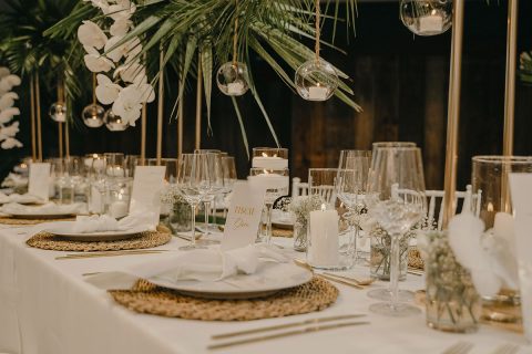 Hochzeitswahn - Elegantes Speisezimmer-Setup für eine Hochzeit mit weiß gedeckten Tischen, Kristallgläsern und goldumrandeten Tellern. Zur Dekoration gehören hängende Glaskugeln und weiße Orchideen vor einem warm beleuchteten Hintergrund. Villa-Flora