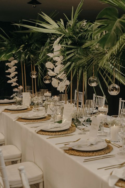 Hochzeitswahn - Elegantes Speisezimmer mit weißen Stühlen und Tischen, geschmückt mit goldenen Tellern, weißen Orchideen, großen Palmblättern und hängenden Glaskugeln. Das Ambiente strahlt eine tropische und doch kultivierte Atmosphäre aus. Villa-Flora