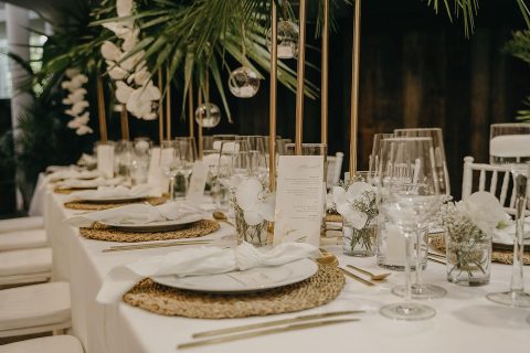 Hochzeitswahn - Eine anspruchsvolle Tischdekoration für den Hochzeitsempfang mit eleganten weißen Blumenarrangements, klarem Glasgeschirr und ordentlich angeordnetem Silberbesteck auf runden gewebten Tischsets, mit einer gut sichtbaren, detaillierten Menükarte. Villa-Flora