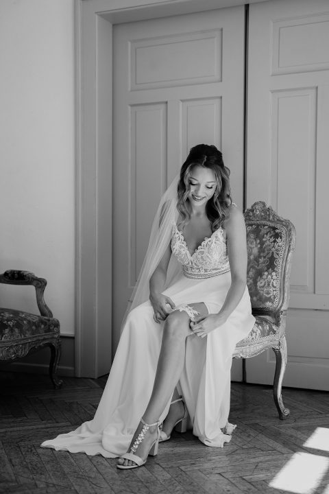 Hochzeitswahn - Eine Braut in einem weißen Kleid und Schleier lächelt, während sie auf einem Stuhl in einem hell erleuchteten Raum mit elegantem Dekor sitzt und ihre Schuhe zurechtrückt. Villa-Flora