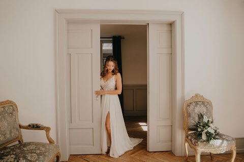 Hochzeitswahn - Eine Frau in einem eleganten weißen Kleid mit Schlitz steht in einem Türrahmen und berührt sanft den Türrahmen. Der Raum ist sonnendurchflutet und mit Sesseln im Vintage-Stil und einem Blumenstrauß ausgestattet. Villa-Flora