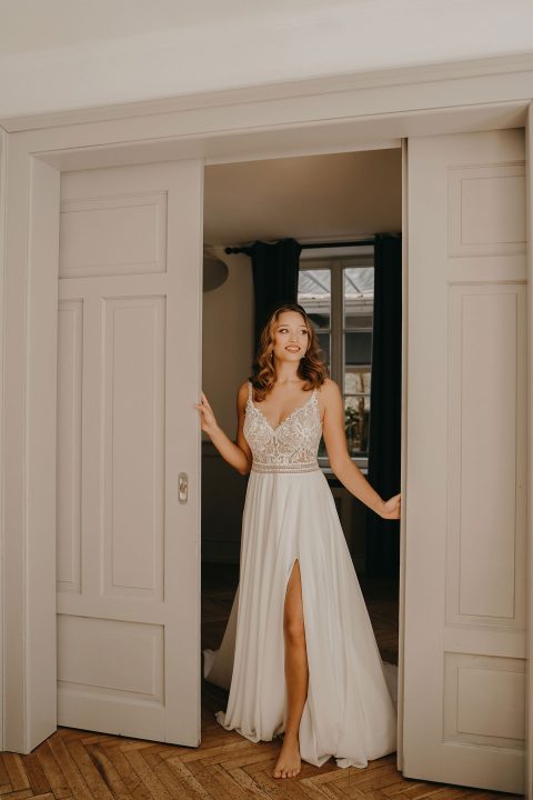 Hochzeitswahn - Eine Frau in einem eleganten weißen Brautkleid mit Spitzendetails und einem oberschenkelhohen Schlitz steht lächelnd in einer Tür in einem Raum mit Holzboden und neutralen Wänden. Villa-Flora