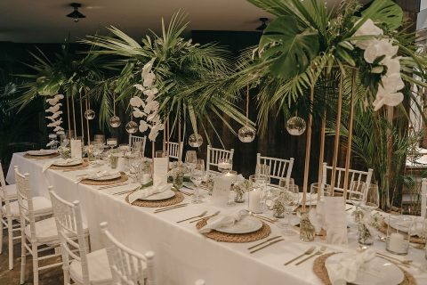 Hochzeitswahn - Elegantes Speisezimmer-Setup für einen besonderen Anlass mit weiß gedeckten Tischen, geschmückt mit goldenen Tellern, Glaswaren und tropischen Blumendekorationen mit großen Palmblättern und hängenden Glaskugeln. Villa-Flora