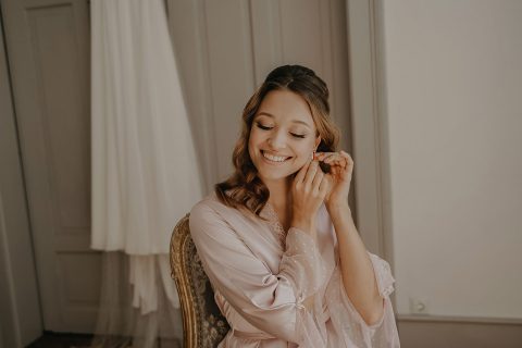 Hochzeitswahn - Eine lächelnde Frau in einem hellrosa Bademantel sitzt in einem sanft beleuchteten Raum mit elegantem Dekor und steckt sich einen Ohrring an. Villa-Flora