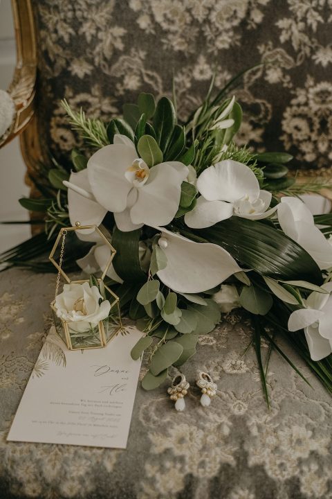 Hochzeitswahn - Eine Hochzeitseinladungskarte liegt neben einem Blumenarrangement mit weißen Orchideen, grünen Blättern und einer kleinen goldenen Laterne auf einem strukturierten grauen Stoff mit einem Vintage-Spiegel im Hintergrund. Villa-Flora
