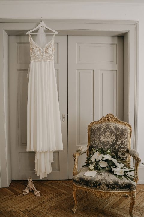Hochzeitswahn - Ein elegantes Brautkleid hängt an einer weißen Tür, dazu Schuhe mit Absatz und ein Blumenstrauß auf einem verzierten Vintage-Stuhl, alles vor einer Wand in neutralen Farben und einem Holzboden. Villa-Flora