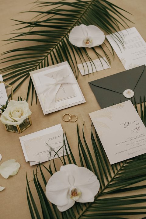 Hochzeitswahn - Hochzeitseinladungspaket auf beigem Hintergrund mit grünen Palmblättern, weißen Orchideen und einer weißen Rose. Enthält Umschläge, Antwortkarten, Haupteinladung mit goldener Schrift und zwei Eheringe. Villa-Flora