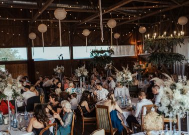 Hochzeitswahn - Eine rustikale und elegant dekorierte Scheune, in der die Gäste an mit Blumendekorationen geschmückten Tischen sitzen und ein festliches Beisammensein unter warmen Lichterketten und weißen Papierlaternen genießen.