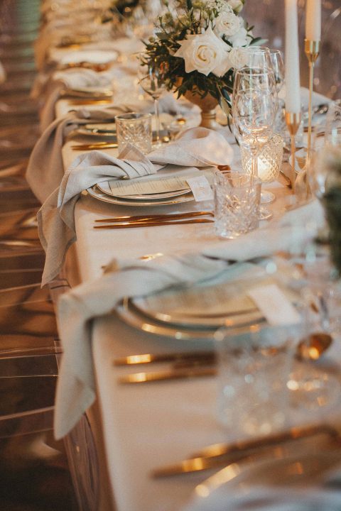 Hochzeitswahn - Ein langer Tisch, gedeckt mit goldenen und weißen Tellern und Besteck.
