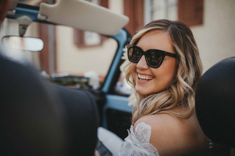 Hochzeitswahn - Eine Braut mit Sonnenbrille sitzt auf dem Rücksitz eines Autos.