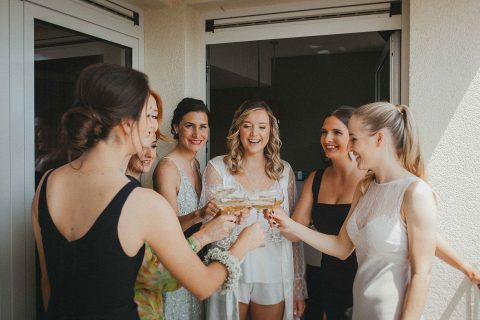 Hochzeitswahn - Eine Gruppe Brautjungfern prostet auf einem Balkon mit Champagner zu.