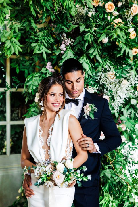 Hochzeitswahn - Eine Braut und ein Bräutigam posieren vor einer Blumenwand.