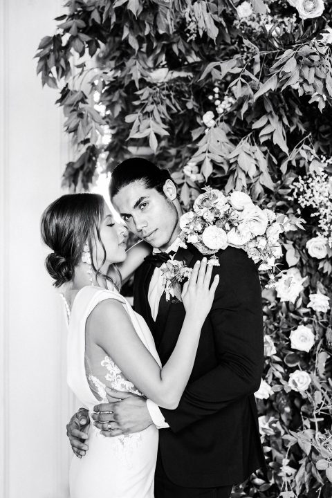 Hochzeitswahn - Ein Schwarz-Weiß-Foto einer Braut und eines Bräutigams, die sich vor einer Blumenwand umarmen.