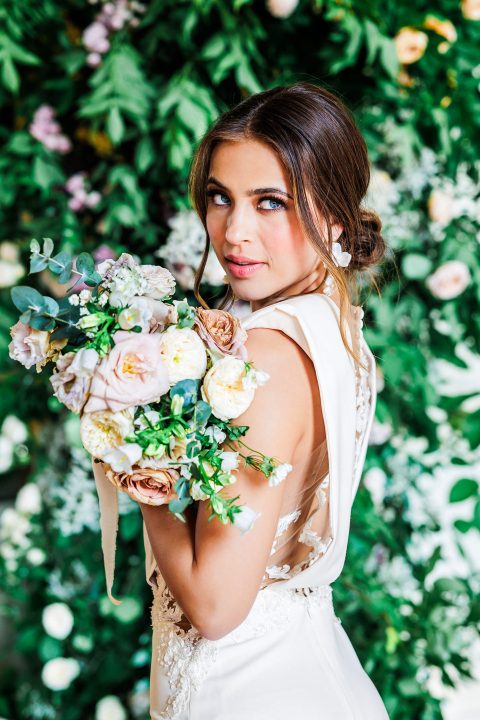 Hochzeitswahn - Eine wunderschöne Braut hält einen Blumenstrauß vor einer grünen Wand.