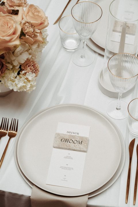 Hochzeitswahn - Ein eleganter Hochzeitstisch mit einer Tischkarte mit der Aufschrift „Bräutigam“, feinem Porzellan, Goldbesteck und Kristallstielen, ergänzt durch ein zartes Blumenarrangement.