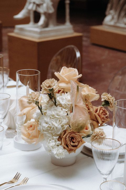 Hochzeitswahn - Eine elegante Tischdekoration mit einem luxuriösen Blumenaufsatz mit zarten Pfirsichrosen, weißen Hortensien und zarten Akzenten, ergänzt durch hohe Glaswaren und feines Silberbesteck, in einem eleganten, kunstvollen Veranstaltungsort.