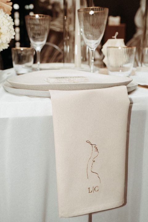Hochzeitswahn - Ein eleganter Tisch mit Kristallglaswaren, einer makellos weißen Tischdecke und einer gefalteten Serviette mit einem gestickten Monogramm „Lic“ darauf wartet auf ein anspruchsvolles kulinarisches Ereignis.