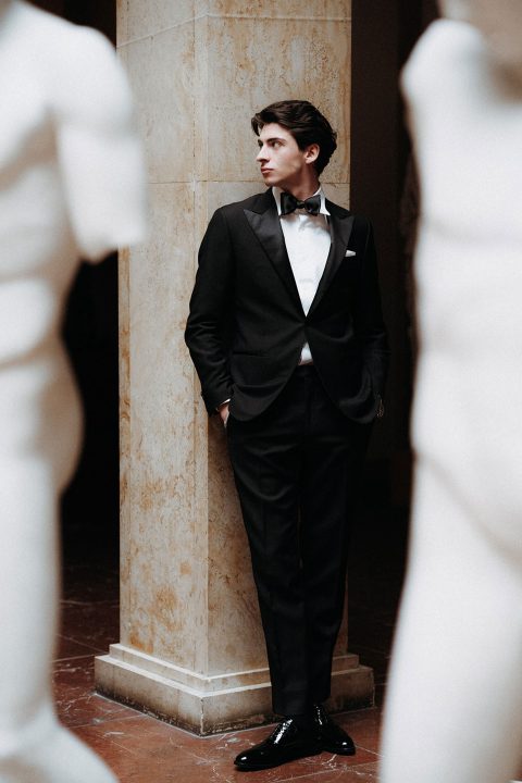 Hochzeitswahn - Ein eleganter junger Mann in einem eleganten schwarzen Smoking und einer Fliege steht nachdenklich zwischen klassischen Statuen und sein nachdenklicher Blick suggeriert einen Moment stiller Selbstbeobachtung inmitten zeitloser Eleganz.