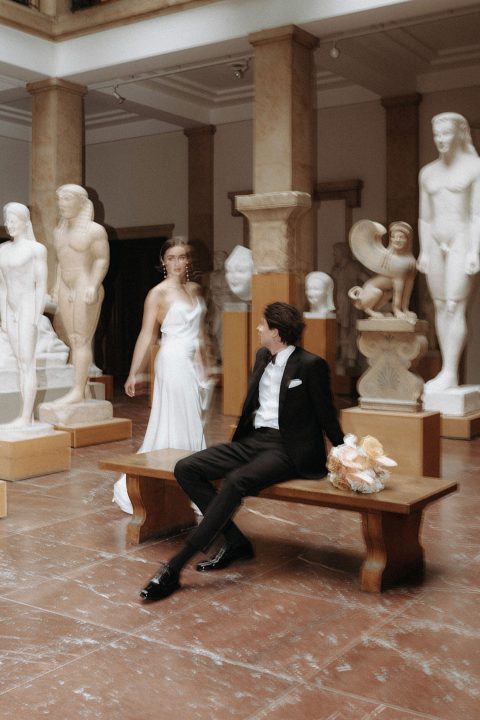 Hochzeitswahn - Eine Braut und ein Bräutigam verbringen einen intimen Moment umgeben von klassischen Statuen in einer Kunstgalerie, die eine Mischung aus zeitgenössischer Romantik und antiker Schönheit verkörpern.