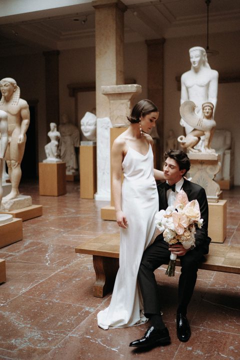 Hochzeitswahn - Ein elegantes Paar posiert nachdenklich in einer klassischen Skulpturengalerie und strahlt inmitten der Marmorfiguren eine zeitlose Romantik aus. Die Frau steht anmutig da, während der Mann sitzt und liebevoll zu ihr aufblickt, beide in formeller Kleidung gekleidet.