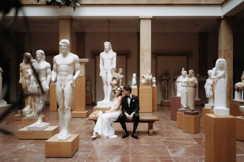 Hochzeitswahn - Ein Paar in eleganter Kleidung sitzt dicht auf einer Bank und tauscht freudige Blicke inmitten einer ruhigen Sammlung klassischer Statuen in einem warm beleuchteten Museumssaal aus und erinnert an eine zeitlose romantische Szene.