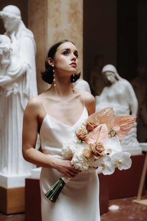 Hochzeitswahn - Eine nachdenkliche Frau in einem eleganten weißen Kleid hält einen Blumenstrauß in der Hand, ihr Blick ist inmitten klassischer Statuen nach oben gerichtet und erinnert an zeitlose Schönheit.