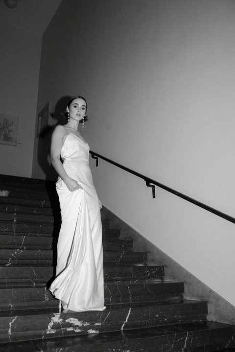 Hochzeitswahn - Eine elegante Frau in einem schicken weißen Kleid bleibt auf einer schwach beleuchteten Treppe stehen. Ihr souveräner Blick kontrastiert mit der schlichten Einfachheit der Umgebung, festgehalten in einem zeitlosen Schwarz-Weiß-Foto.