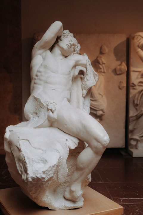 Hochzeitswahn - Eine klassische Marmorskulptur zeigt eine sitzende männliche Figur in nachdenklicher Pose, deren Muskulatur detailliert und der Ausdruck nachdenklich ist, vor dem Hintergrund anderer Skulpturen in einer Galerieumgebung.