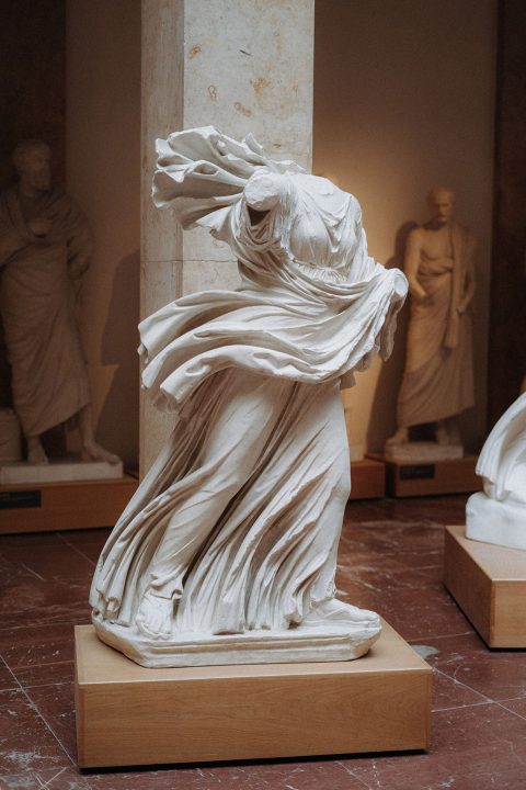Hochzeitswahn - Eine Marmorstatue, die eine drapierte weibliche Figur darstellt, möglicherweise aus der klassischen Antike, mit komplizierten Falten der Kleidung, ausgestellt in einer Museumsgalerie mit anderen Skulpturen im Hintergrund.