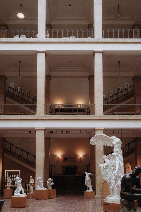 Hochzeitswahn - Eine elegante, geräumige Museumshalle, geschmückt mit klassischen Skulpturen, mit großen Säulen und mehreren Ebenen, die durch Treppen verbunden sind und von einem warmen, stimmungsvollen Licht beleuchtet werden.
