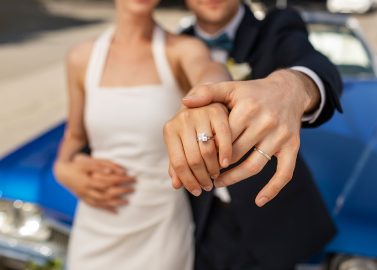 Hochzeitswahn - Ein Paar in formeller Kleidung hält den perfekten Moment fest, indem es seine Trauzeugen vor einem blauen Auto präsentiert. Die Frau präsentiert in einem weißen Kleid elegant ihren Verlobungsring und der Mann steht neben ihr und zeigt stolz seinen Ehering. Zuckerbergschloss-Kappelrodeck-Hochzeit
