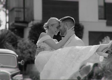 Hochzeitswahn - Draußen hebt ein Bräutigam im Anzug die Braut im weißen Brautkleid hoch und umarmt sie. Beide lächeln und berühren ihre Stirn. Im Hintergrund sind ein Oldtimer und ein modernes Haus zu sehen, die einen freudigen Moment einfangen – gestaltet von McKenzie Productions. Zuckerbergschloss-Kappelrodeck-Hochzeit