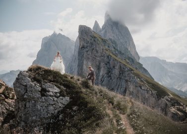 Hochzeitswahn - Eine Braut im weißen Kleid und ein Bräutigam im Anzug stehen auf einem grasbewachsenen, felsigen Berghang mit dramatischen, zerklüfteten Gipfeln und Wolken im Hintergrund, wunderschön eingefangen von Francesco Calarota Photography. Zuckerbergschloss-Kappelrodeck-Hochzeit