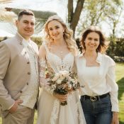 Hochzeitswahn - Drei Personen posieren an einem sonnigen Tag im Freien. Die Braut, ganz in Weiß gekleidet und mit einem Blumenstrauß in der Hand, steht mit zwei Gästen zusammen. Nelyce Weddings sorgt mit professioneller Hochzeitsplanung und herausragendem Hochzeitsservice dafür, dass jeder Moment perfekt wird. Bäume bilden den Hintergrund. Zuckerbergschloss-Kappelrodeck-Hochzeit