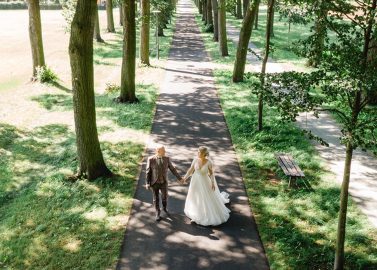 Hochzeitswahn - An einem sonnigen Tag gehen Braut und Bräutigam Hand in Hand einen von Bäumen gesäumten Weg in einem Park entlang. Eingefangen von Jens Schaffhirt Photography, erzeugen die Schatten der Bäume beim Spazierengehen kunstvolle Muster auf dem Weg.